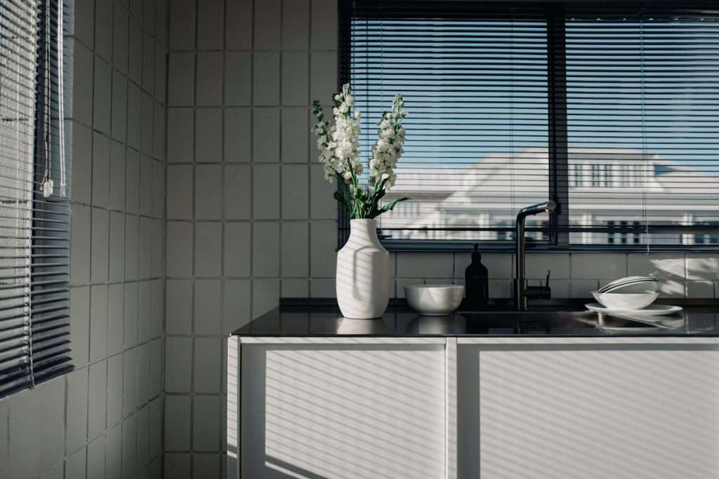 a white vase with flowers on a kitchen counter near clean windowsill